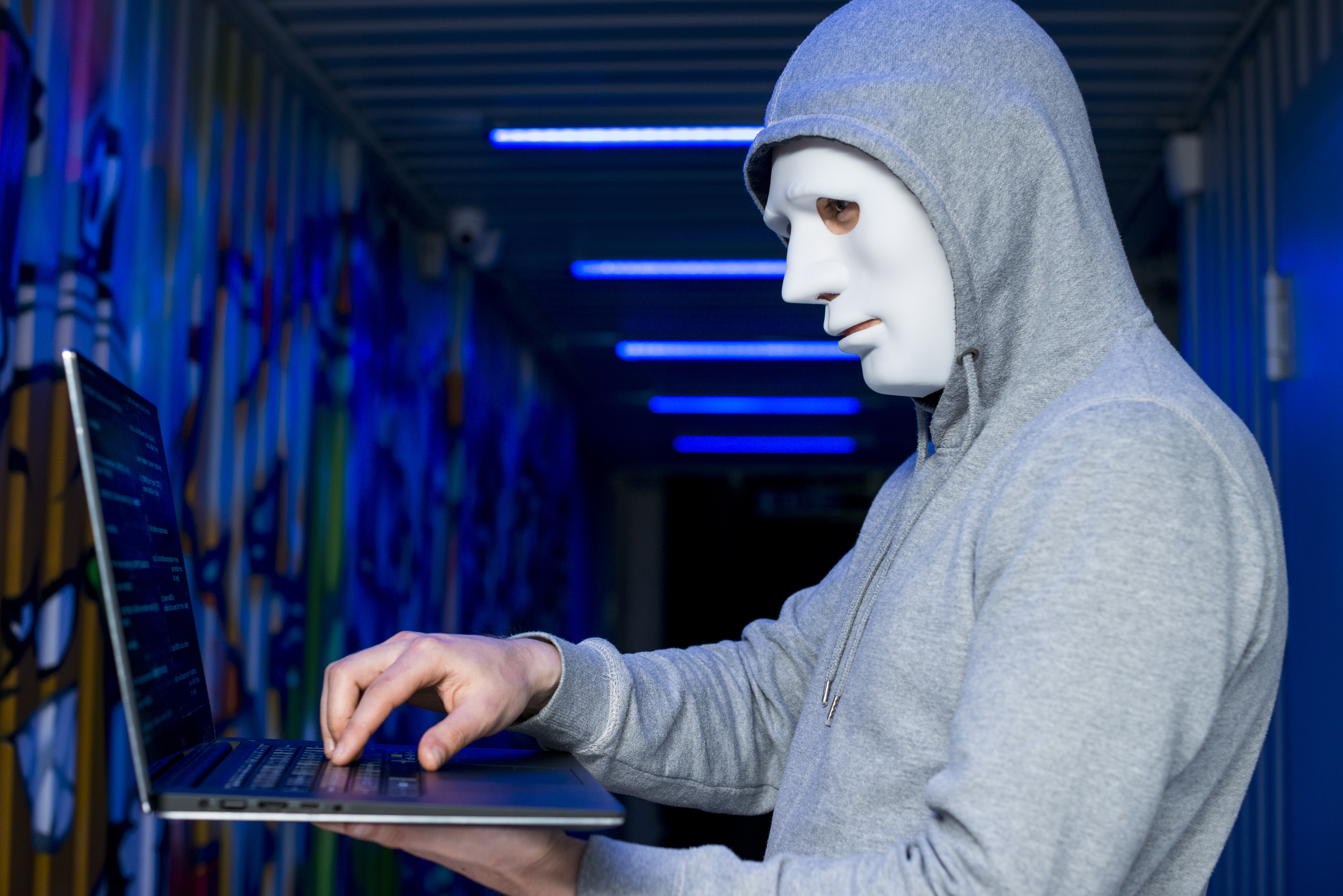 Sosok anonim bertopeng memakai hoodie abu-abu sedang menggunakan laptop, menggambarkan peretas yang mengambil alih domain PeduliLindungi dalam serangan siber.